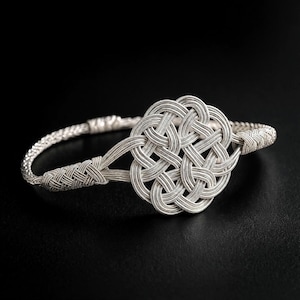 May include: A silver bracelet with a Celtic knot design. The bracelet features a woven band and a central knot element, all in a bright silver color. The intricate knotwork is the focal point, set against a dark background.