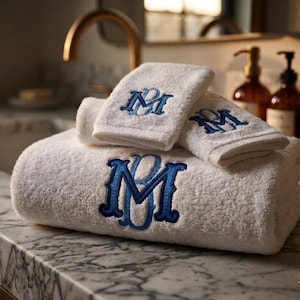 May include: A set of white terry cloth towels featuring a blue embroidered monogram with the letters BM. The towels are stacked, with the largest one on the bottom and two smaller towels folded on top, all displaying the same blue lettering.