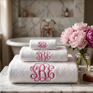 May include: A stack of three white terry cloth towels, each featuring a pink embroidered monogram with the letters SBE in a decorative script font. The towels are arranged from smallest to largest, with the monogram centered on each piece.