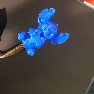 May include: A vibrant blue figurine of a cartoon character, 3D-printed and attached to a gold-coloured metal object. The character is lying down. The metal object has the letters "SC" engraved on it.