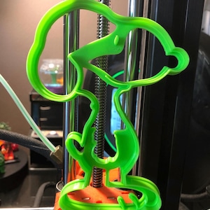 Pode incluir: Uma figura verde brilhante impressa em 3D de Snoopy, o beagle de desenho animado, montada em uma base laranja. A figura está presa a uma haste de metal, provavelmente parte de uma impressora 3D. O design é uma representação lúdica do personagem.