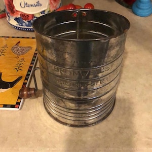 Pode incluir: Um peneira de medição vintage de metal prateado com uma manivela de madeira. A peneira tem as palavras "Bromwell's Measuring Sifter" impressas no lado.