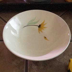 Puede incluir: Cuenco de cerámica blanquecina con un diseño floral amarillo y verde. El cuenco presenta una flor amarilla estilizada con hojas verdes y un pequeño pétalo amarillo. El cuenco tiene forma redondeada y acabado brillante.