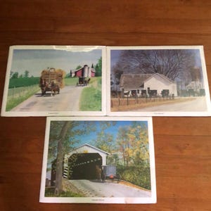 May include: Three framed landscape prints featuring rural scenes. One print shows a horse-drawn carriage on a road, another depicts a white farmhouse, and the third showcases a covered bridge. The prints have a vintage aesthetic.