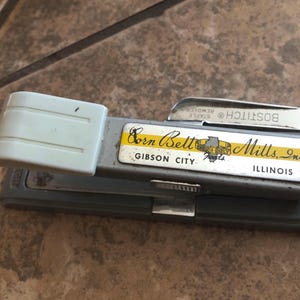 May include: A vintage gray stapler with a light blue handle and a silver staple remover. The stapler has a label that reads "Corn Belt Mills, Inc. GIBSON CITY ILLINOIS". The staple remover is marked "BOSTITCH STAPLE REMOVER".