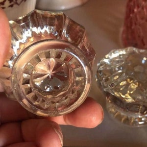 May include: Two clear glass bottle stoppers with intricate designs. One stopper has a star-shaped design in the center, while the other has a more floral pattern.