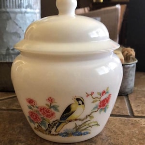 Op de afbeelding: Een witte keramische pot met een deksel, versierd met een geschilderd ontwerp van een gele vogel op een tak met rode en roze bloemen. De pot heeft een afgeronde vorm en een decoratieve knop op het deksel.