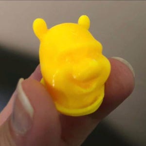 Puede incluir: Una figura de plástico amarillo brillante de la cabeza de un oso de dibujos animados, con orejas pequeñas y redondeadas y una expresión sonriente. La figura se sostiene en la mano de una persona, con la cara del oso claramente enfocada.