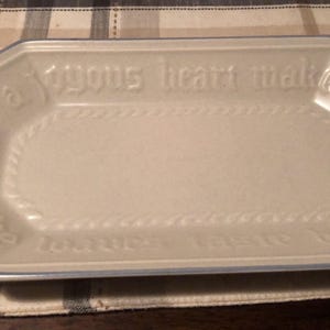 May include: A rectangular, off-white ceramic serving platter with a blue trim. The platter has the embossed text "A joyous heart makes the loaves taste better" and a decorative rope-like border.