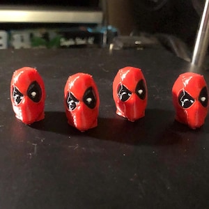 Puede incluir: Cuatro pequeñas cabezas rojas de Deadpool con detalles de ojos negros están dispuestas en fila. Las cabezas están impresas en 3D y reposan sobre una superficie oscura. El diseño icónico de la máscara es claramente visible.