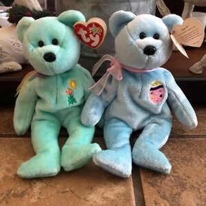 May include: Two plush teddy bears in shades of blue and teal. The teal bear has a floral design and the blue bear has an Easter egg design. Both have black eyes, black noses, and attached tags.