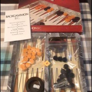May include: A vintage backgammon game set with a gray and white game board, orange and black playing pieces, and two dice. The game board is in a plastic case with a clear plastic cover. The box is labeled "Backgammon B.C. The ancient game made new."