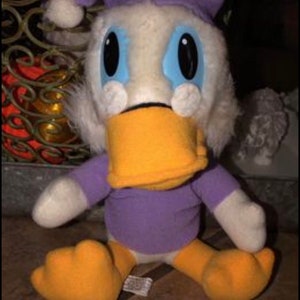 May include: A plush toy of a cartoon duck wearing a purple nightcap and shirt, with an orange beak and feet. The duck has large blue eyes and white fur around its face. The toy is sitting on a brown surface.