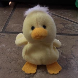 Könnte beinhalten: Ein gelbes Plüsch-Entenspielzeug mit orangefarbenem Schnabel und Füßen. Die Ente hat schwarze Augen und einen Büschel weißen Fluffs auf dem Kopf. Das Spielzeug ist als kleines Entenküken gestaltet.