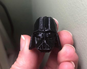 May include: A black 3D-printed Darth Vader helmet, a recognizable character from the Star Wars franchise. The helmet is detailed with the iconic mask and helmet design. The object is held in a person's hand.