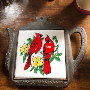 May include: A cast iron trivet shaped like a teapot with a square ceramic tile insert. The tile features a colourful illustration of two bright red cardinal birds perched on a green branch with yellow flowers against a white background.