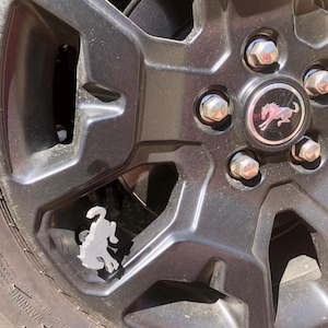 May include: A matte black vehicle wheel featuring a central hub cap with a silver running horse emblem. A white, three-dimensional running horse figure is attached to the valve stem, contrasting against the dark finish of the wheel rim.