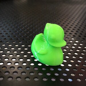 May include: A bright green 3D printed rubber duck toy. The duck is sitting on a black surface with a grid pattern.