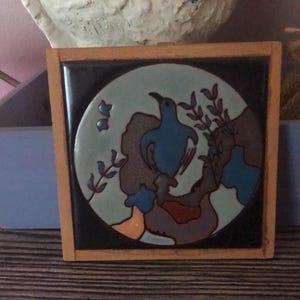 Może przedstawiać: Kwadratowa płytka ceramiczna z motywem ptaka w drewnianej ramie. Ptak jest niebieski, na jasnoniebieskim tle z brązowymi i niebieskimi akcentami. Rama wykonana jest z jasnego drewna.