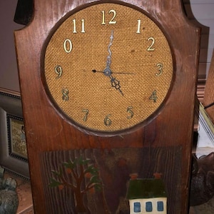 May include: A wooden wall clock with a burlap face and gold numbers. The clock has a painted scene of a house, a tree, and a fence below the clock face.
