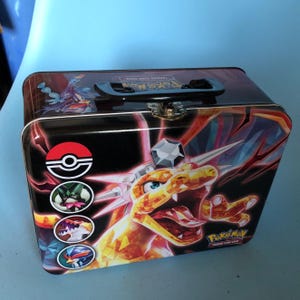 Puede incluir: Una lata de colección rectangular de metal para el juego de cartas coleccionables Pokémon, con fondo negro y una criatura tipo dragón naranja-amarilla facetada. Incluye cuatro iconos circulares, el logo de Pokémon y un asa de plástico negra.
