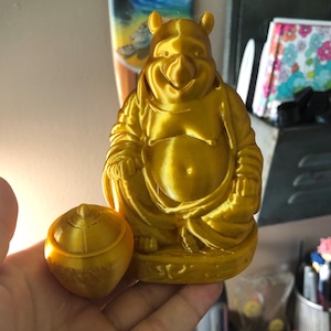 Puede incluir: Una figura dorada de un personaje sonriente en posición sentada, con un pequeño recipiente redondo y tapado al lado. La figura tiene una gran barriga y orejas distintivas. Probablemente sea un objeto decorativo o de colección.