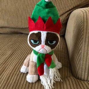 May include: A plush toy of Grumpy Cat wearing a green elf hat with a white pom-pom and a red crown. The cat has a green and red scarf with white fringe. The cat has a grumpy expression.