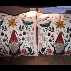 May include: Two white cushion covers with a red, green and gold festive design featuring a gnome, birds and snowflakes.