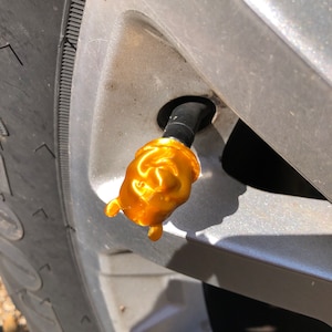 Puede incluir: Un tapón de válvula de neumático de color dorado con forma de oso de dibujos animados. El oso tiene una cabeza redonda, orejas pequeñas y una nariz pequeña.