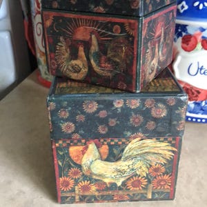 May include: Two decorative boxes stacked on top of each other. The boxes are black with a floral pattern and rooster illustrations. The rooster illustrations are yellow, orange, and brown. The boxes are likely for storage or decoration.