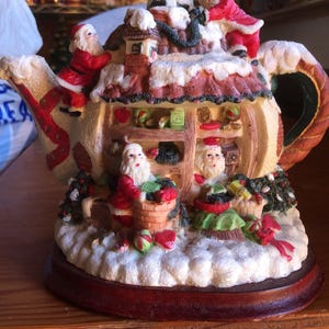 Puede incluir: Una figura decorativa de tetera navideña con figuras de Papá Noel y una escena de pueblo nevado. La tetera es de color crema con detalles en rojo y verde, y se asienta sobre una base de madera marrón oscuro. La figura mide aproximadamente 20 cm de alto.