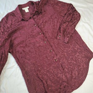 May include: A long-sleeved burgundy button-down shirt with a floral pattern. The shirt has a collar and a button-up front.