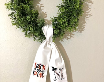 May include: White fabric bow with the text "trick or treat" and a witch's hat on one side and the letter "M" with a witch's hat on the other side. The bow is tied around a green wreath.