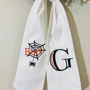 Puede incluir: Lazo de tela blanco con una telaraña negra y la palabra "Boo!" bordada en un lado y la letra "G" bordada en el otro.