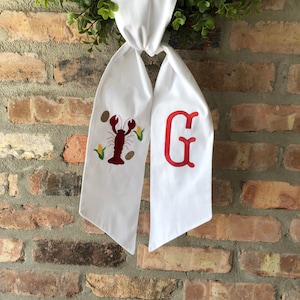 May include: White fabric sash with a red embroidered lobster and corn on one side and a red embroidered letter G on the other side.