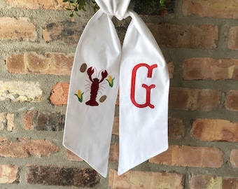 Op de afbeelding: Witte stoffen sjerp met een rood geborduurde kreeft en maïs aan de ene kant en een rood geborduurde letter G aan de andere kant.