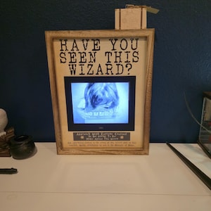 May include: A rectangular wooden frame containing a digital screen displaying a black and white image of a person holding a sign. The frame features the text HAVE YOU SEEN THIS WIZARD? at the top and warning text at the bottom of the screen.