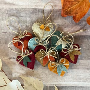 K&ouml;nnte beinhalten: Eine Sammlung von herzf&ouml;rmigen Filzanh&auml;ngern in verschiedenen Herbstfarben wie Dunkelrot, gebranntem Orange, Senfgelb und gedecktem Blaugr&uuml;n. Jedes Herz ist mit einer kleinen Juteschleife und zwei Filzbl&auml;ttern an einer Schnur zum Aufh&auml;ngen versehen.