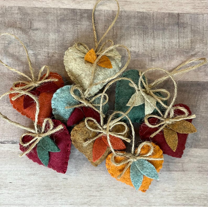 K&ouml;nnte beinhalten: Eine Sammlung von sieben herzf&ouml;rmigen Filzanh&auml;ngern in verschiedenen Farben wie Rot, Orange, Gelb, T&uuml;rkis und Gr&uuml;n. Jedes Herz ist mit einem kleinen Filzblatt-Akzent und einer Schlaufe aus Jutekordel versehen, die oben zu einer Schleife gebunden ist.