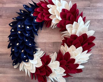 Könnte beinhalten: Ein patriotischer Kranz im Stil der amerikanischen Flagge. Er hat abwechselnde Abschnitte aus roten, weißen und blauen Filzblüten. Der blaue Abschnitt hat weiße Sternakzente und schafft ein festliches und dekoratives Stück.