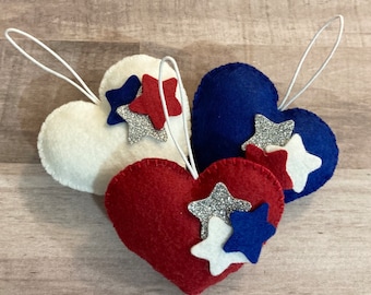 May include: Three felt heart ornaments with stars. The hearts are red, white, and blue. The stars are silver, white, and blue.