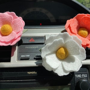 May include: Three felt flower air fresheners in a car. The flowers are pink, white, and coral with yellow centers. The flowers are hanging from the air vents in the car.
