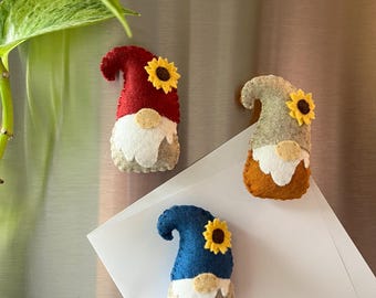 Könnte beinhalten: Drei Filz-Zwerg-Magnete in Rot, Blau und Beige, jeweils mit Sonnenblumenakzent. Die Zwerge haben weiße Bärte und beige Körper. Sie werden auf einer Metalloberfläche mit weißem Papier angezeigt.