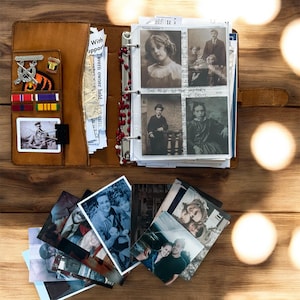 May include: A brown leather journal with a ring binder, open to display vintage photographs. The journal has a pocket with military pins and ribbons. Loose photos are scattered around the journal on a wooden surface. The journal is a keepsake.