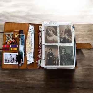 May include: Open brown leather journal with a strap closure, filled with vintage photographs, military pins, and ribbons. The journal's interior includes a rosary and various documents. The photographs are black and white, depicting people from the past.