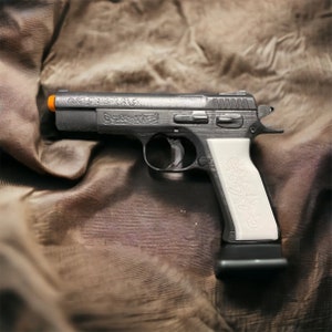 Peut inclure: Un pistolet argenté avec une poignée blanche ornée d'un motif floral complexe. L'arme repose sur une surface en tissu brun.