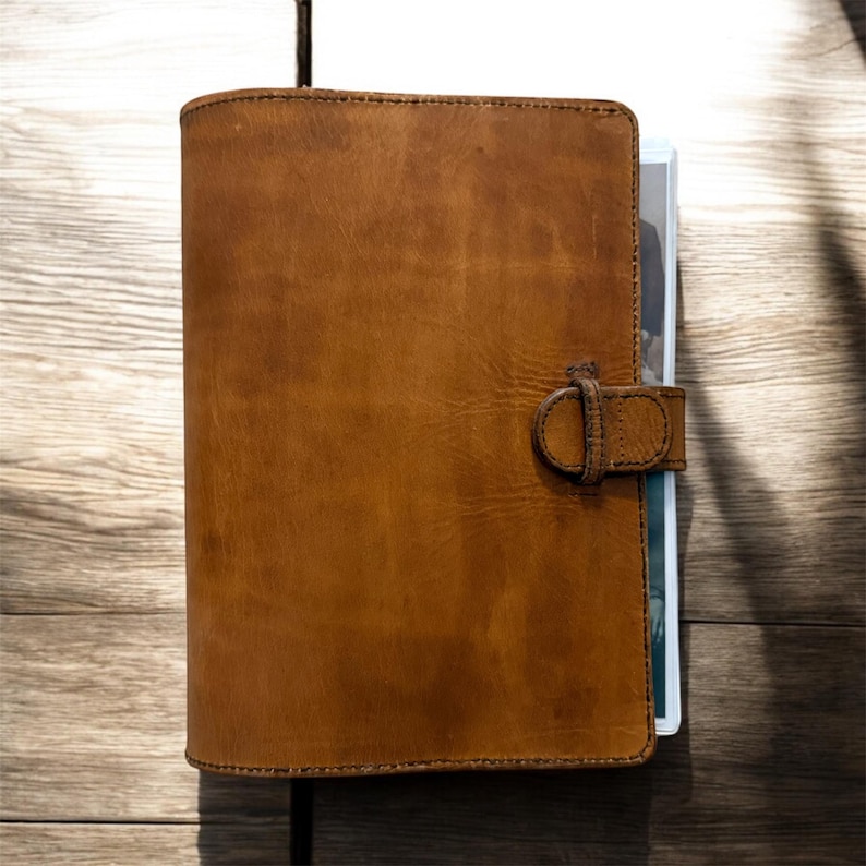 May include: A brown leather journal with a buckle closure. The journal is closed and appears to be filled with pages. The leather has a textured appearance and is a warm, medium brown color. The journal is resting on a wooden surface.