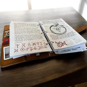 May include: Open brown leather journal with handwritten notes and illustrations. The journal is open to a page with text and symbols in red ink. A metal ring binder holds the pages together. Keys and a card are visible in a side pocket.
