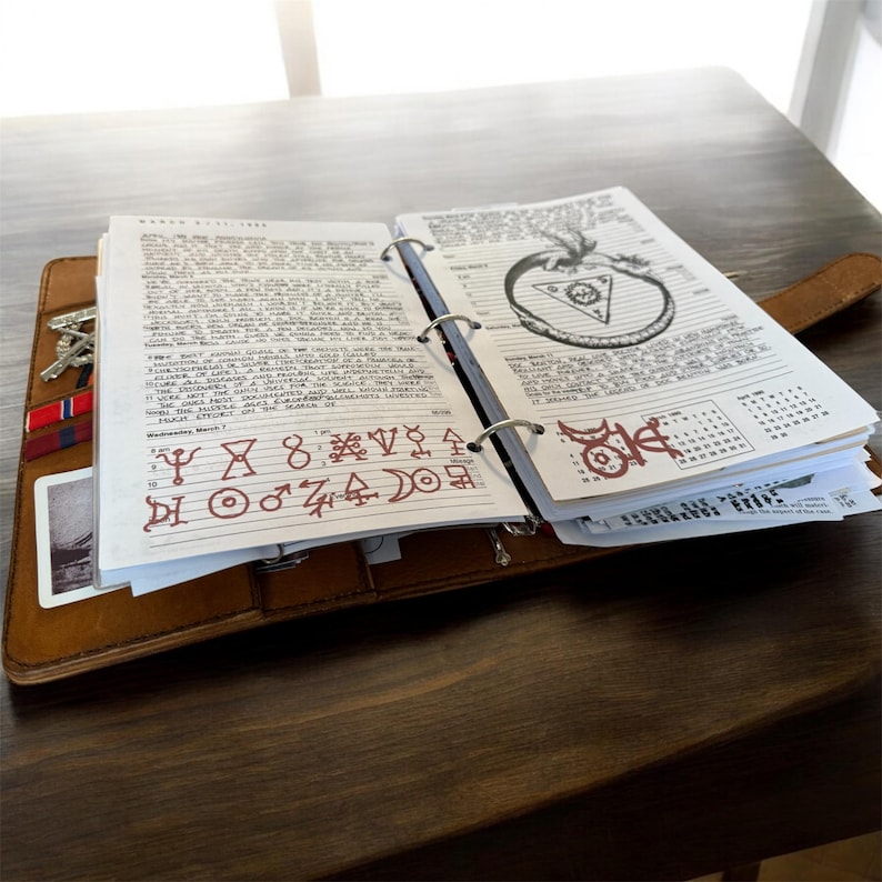May include: Open brown leather journal with handwritten notes and illustrations. The journal is open to a page with text and symbols in red ink. A metal ring binder holds the pages together. Keys and a card are visible in a side pocket.