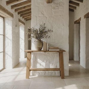 Puede incluir: Una mesa consola de madera de color marrón claro con un estante inferior, coronada con un gran jarrón de cerámica desgastada que contiene ramas de olivo verdes y una pequeña pila de tres libros, colocada contra una pared de piedra blanca.